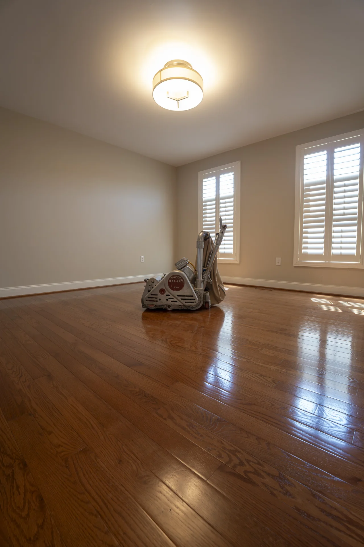 Empty room with a floor sander, showing newly refinished, glossy brown hardwood floors. by Tesla Hardfloor in Doylestown, PA