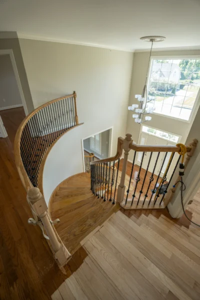 High-angle view of a grand wooden spiral staircase during refinishing, with light wood and dark wood sections. by Tesla Hardf
