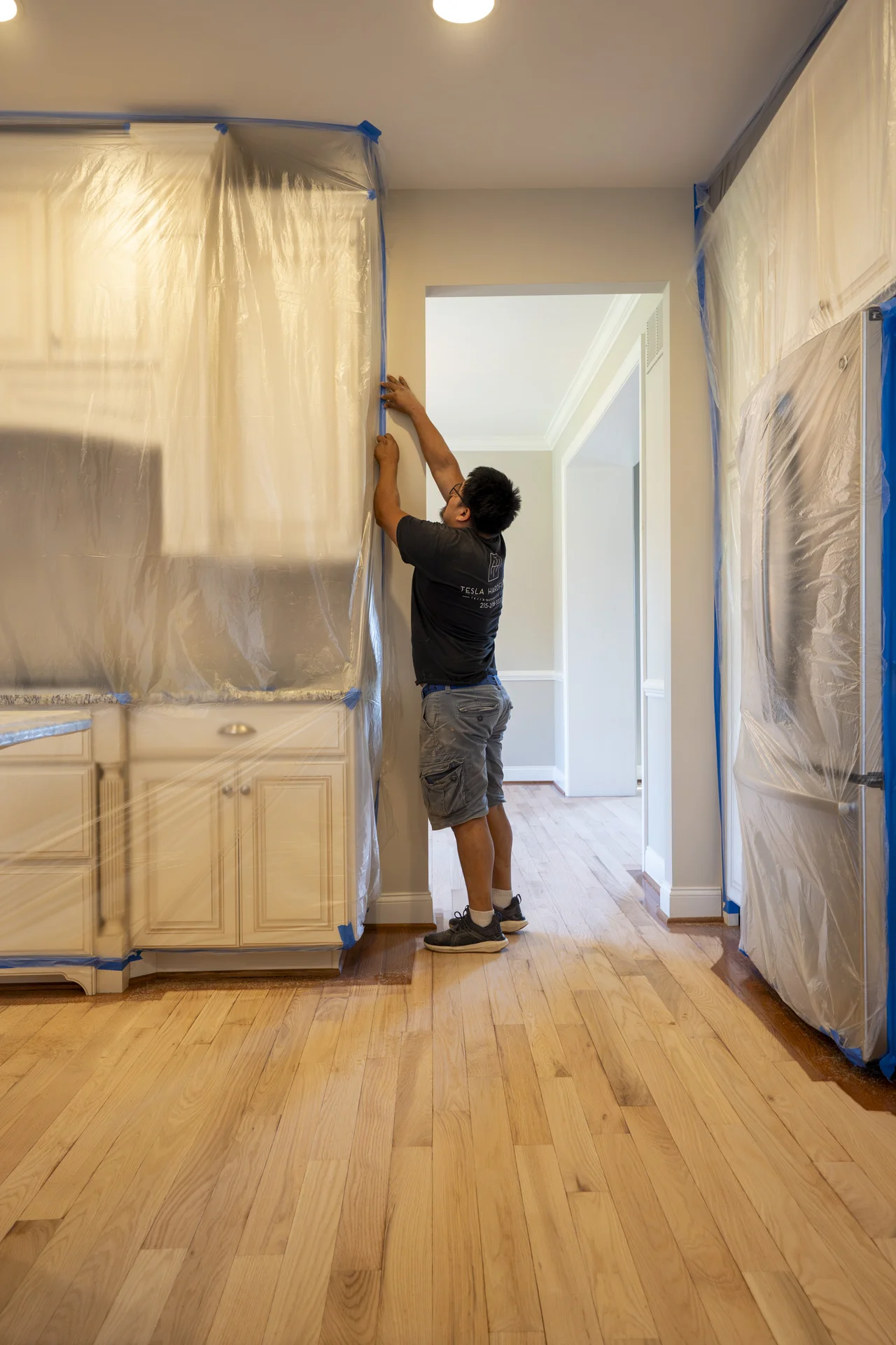 Worker taping plastic sheeting to protect kitchen cabinets during hardwood floor refinishing. by Tesla Hardfloor in Doylestown
