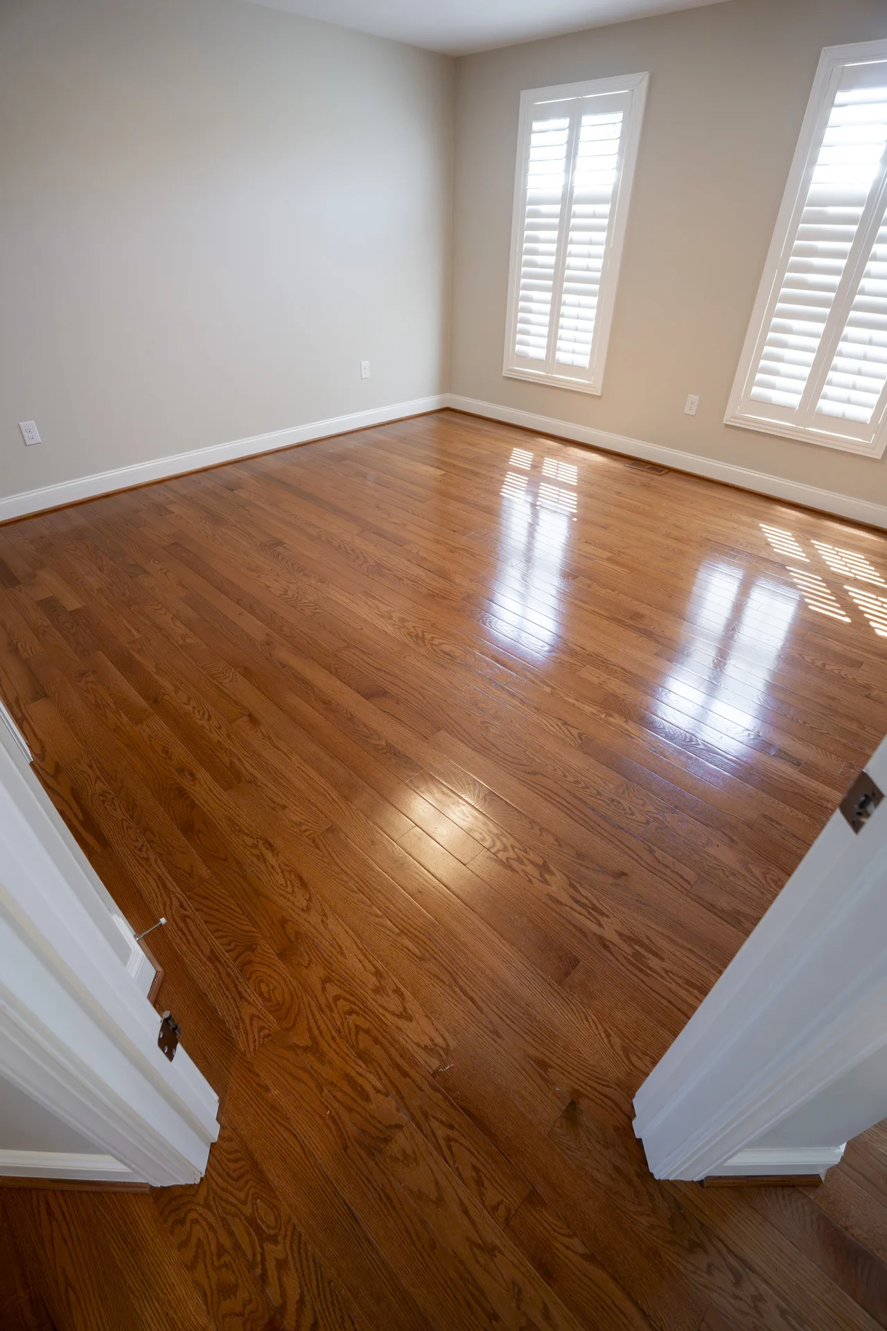 Custom Hardwood Floor Design in Newtown, PA Polished medium-brown hardwood flooring installed in an empty room with white baseboards and window shutters. by Tesla Hardfl